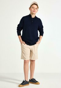 Marinblå zip-tröja, ljusbeiga shorts och svarta sneakers med gummisula. Tröjan har lösa ärmar och en avslappnad passform.
