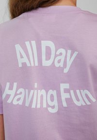 Paarse t-shirt met een speelse witte tekstontwerp op de achterkant dat "De hele dag plezier hebben" leest. Zachte stof, korte mouwen.