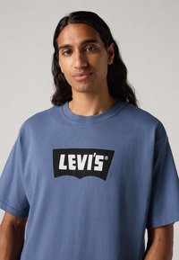 Mėlyna medvilninė marškinėliai su juodu Levi's logotipu kreivame dizaine priekyje. Trumpomis rankovėmis ir klasikiniu apykaklės modeliu.