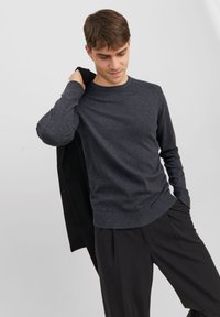 Un pull ras du cou gris foncé en tissu doux et texturé. Présente des poignets et un ourlet côtelés. Le modèle tient un blazer noir sur une épaule.