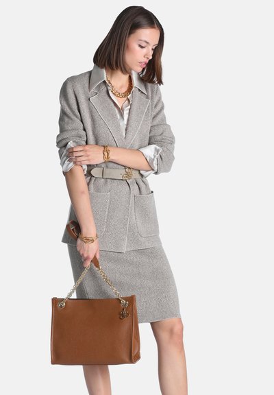 Femme en tailleur jupe en chevron gris avec une ceinture beige, tenant un sac à main en cuir marron avec des poignées en chaîne dorée.