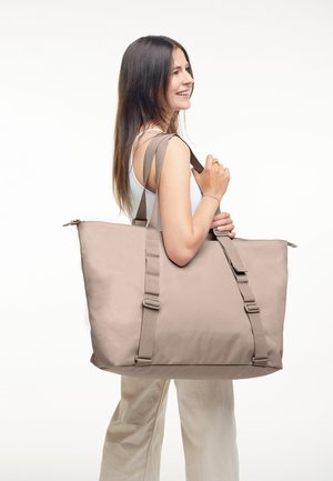 Donna che porta una grande borsa a tracolla beige con cinturini regolabili, sorridente, indossa una canotta bianca e pantaloni chiari su uno sfondo semplice.