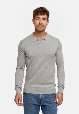 Unge mand med krøllet hår iført en lysegrå langærmet polo sweater og blå jeans, stående mod en ensfarvet hvid baggrund.