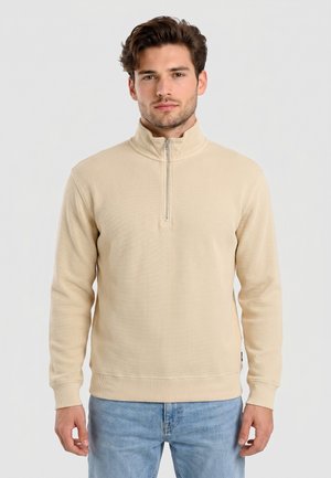 Giovane uomo con capelli corti castani che indossa un maglione beige con zip a quarto e jeans azzurri, in piedi davanti a uno sfondo semplice.