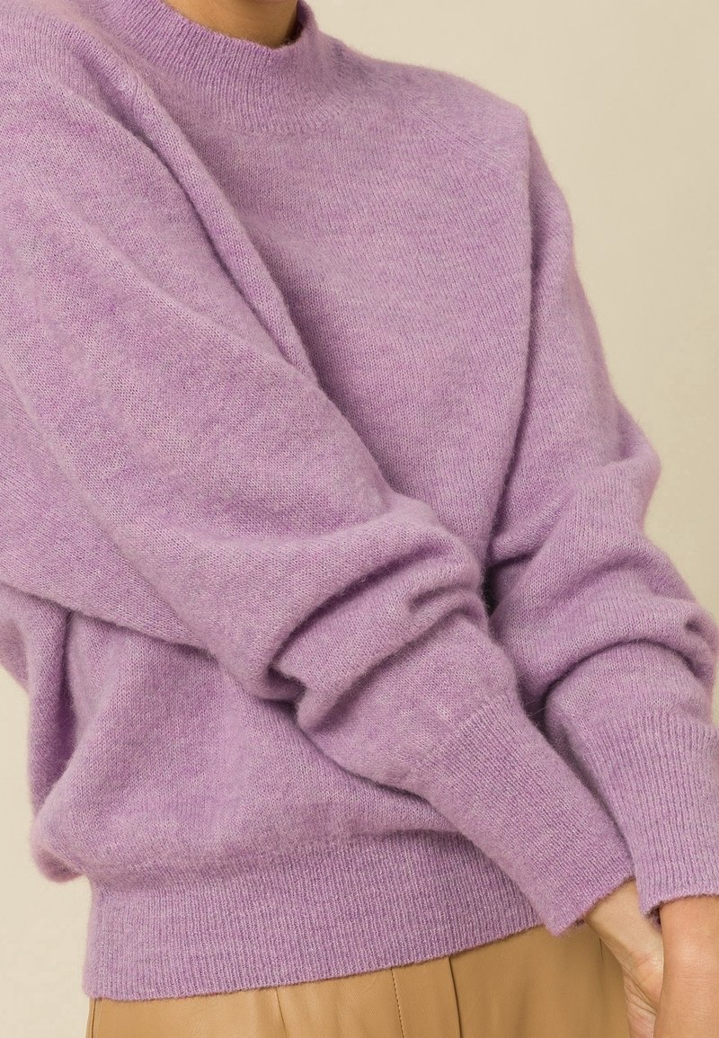 Personne portant un pull doux lavande à manches longues retroussées aux poignets, associé à un pantalon marron clair, sur un fond neutre.