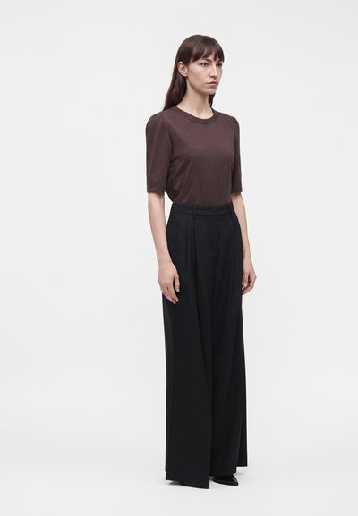 Femme debout portant un haut marron à manches courtes et un pantalon noir taille haute à jambes larges sur un fond clair uni.