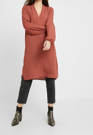 Frau trägt ein rostfarbenes, strukturiertes Langarmkleid über schwarzen, gekürzten Jeans und Schlangenleder-Stiefeletten, steht vor einem schlichten Hintergrund.