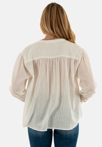 Blusa blanca ligera con mangas largas, detalle plisado en la parte posterior y una textura de rayas sutil, combinada con jeans azules.