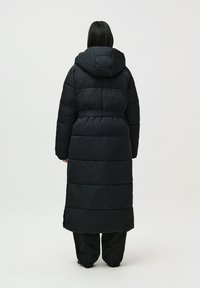 Mujer de pie de espaldas, vistiendo un abrigo largo negro acolchado con capucha y cinturón, junto con pantalones anchos negros, frente a un fondo claro y liso.