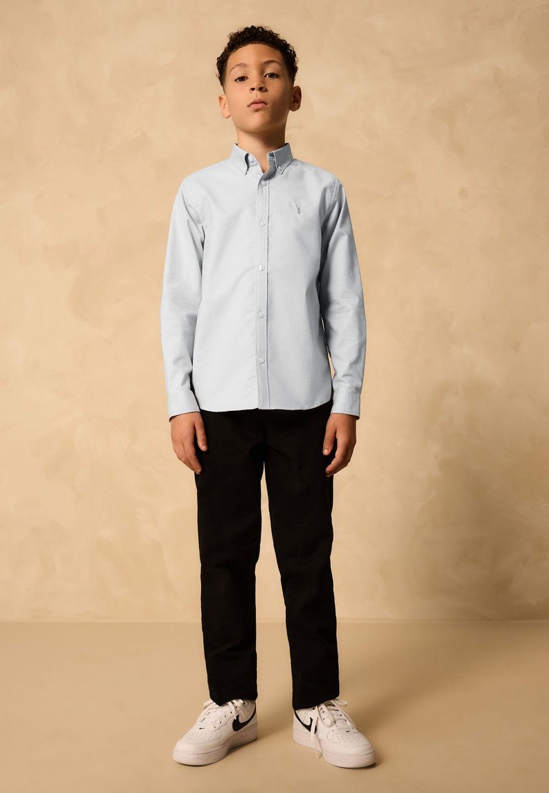 Garçon debout portant une chemise bleu clair à boutons, un pantalon noir et des baskets blanches Nike sur un fond beige uni.
