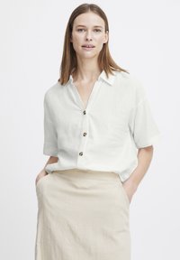 Camicia bianca in lino a maniche corte con colletto, caratterizzata da bottoni di un colore a contrasto, indossata con una gonna beige chiara.