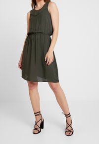 Olivgrön ärmlös klänning med en samlad midja och rund halsringning. Kombinerad med svarta högklackade sandaler med remmar.