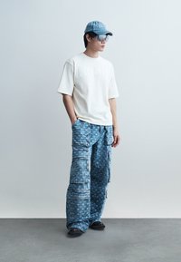 Mladý muž stojí oblečený v bielom nadrozmernom tričku, modrých cargo nohaviciach s vzorom, čiernych topánkach, modrom denimovom klobúku a veľkých slnečných okuliaroch.