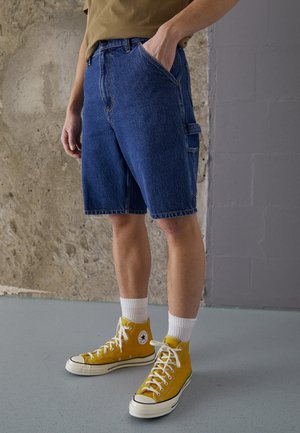 Personne portant un short en jean bleu, des baskets montantes jaunes, des chaussettes blanches et une chemise marron, debout contre un mur texturé à l'intérieur.