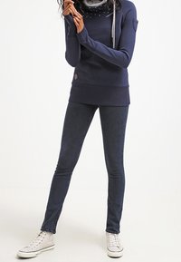 Marinblå långärmad tröja med texturerad tyg, silverfärgade detaljer och en logotypapplikation, kombinerad med mörka skinny jeans och vita sneakers.