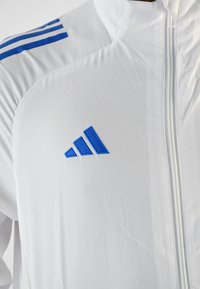 Chaqueta deportiva blanca con un acabado texturizado, rayas horizontales azules en los hombros y un logo bordado azul en el pecho.