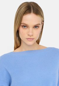 Maglione azzurro a coste con scollo ampio. Il modello ha capelli lunghi e lisci e orecchini a catena. Design semplice e moderno.