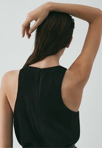 Mustassa hihattomassa vaatteessa on sileä pinta ja korkea kaula-aukko. Siinä on huomaamaton vetoketju takana ja se laskeutuu rennosti olkien yli.