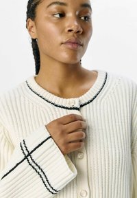 Cardigan crema a coste con rifiniture nere, scollo rotondo e bottoni in perla. Caratterizzato da maniche larghe e un design a maglia testurizzato.
