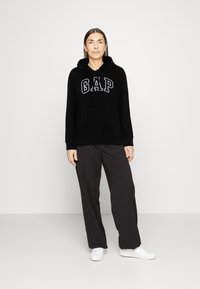 Svart hoodie med "GAP"-logotyp i grått, med en känguruficka och dragsko i huvan, tillsammans med löst sittande mörka byxor och vita sneakers.