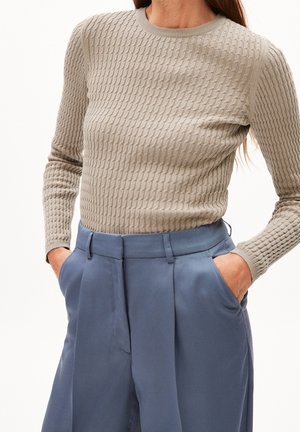 Femme portant un pull beige texturé à manches longues, rentré dans un pantalon bleu taille haute, les mains dans les poches.