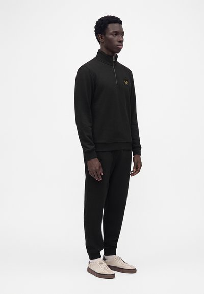 Jeune homme debout, portant un pull noir zippé et un pantalon noir avec des baskets beige sur un fond blanc uni.