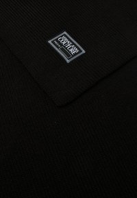 Sort strikket stof med en ribbet tekstur, der har et rektangulært mærke, som der står "VERSACE JEANS COUTURE" på.