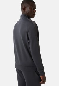 Ciemnoszara sportowa kurtka z zamkiem błyskawicznym i wysokim kołnierzem, z prążkowanymi mankietami i dołem. Gładka tekstura z subtelnymi akcentami po bokach. Stylizowana na odzież codzienną dla aktywnych.