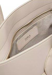 Beige leren handtas interieur met een zachte textuur, voorzien van een ritsvak en een label met de tekst "FURLA SINCE 1927" in reliëf. Gouden ritsdetail.