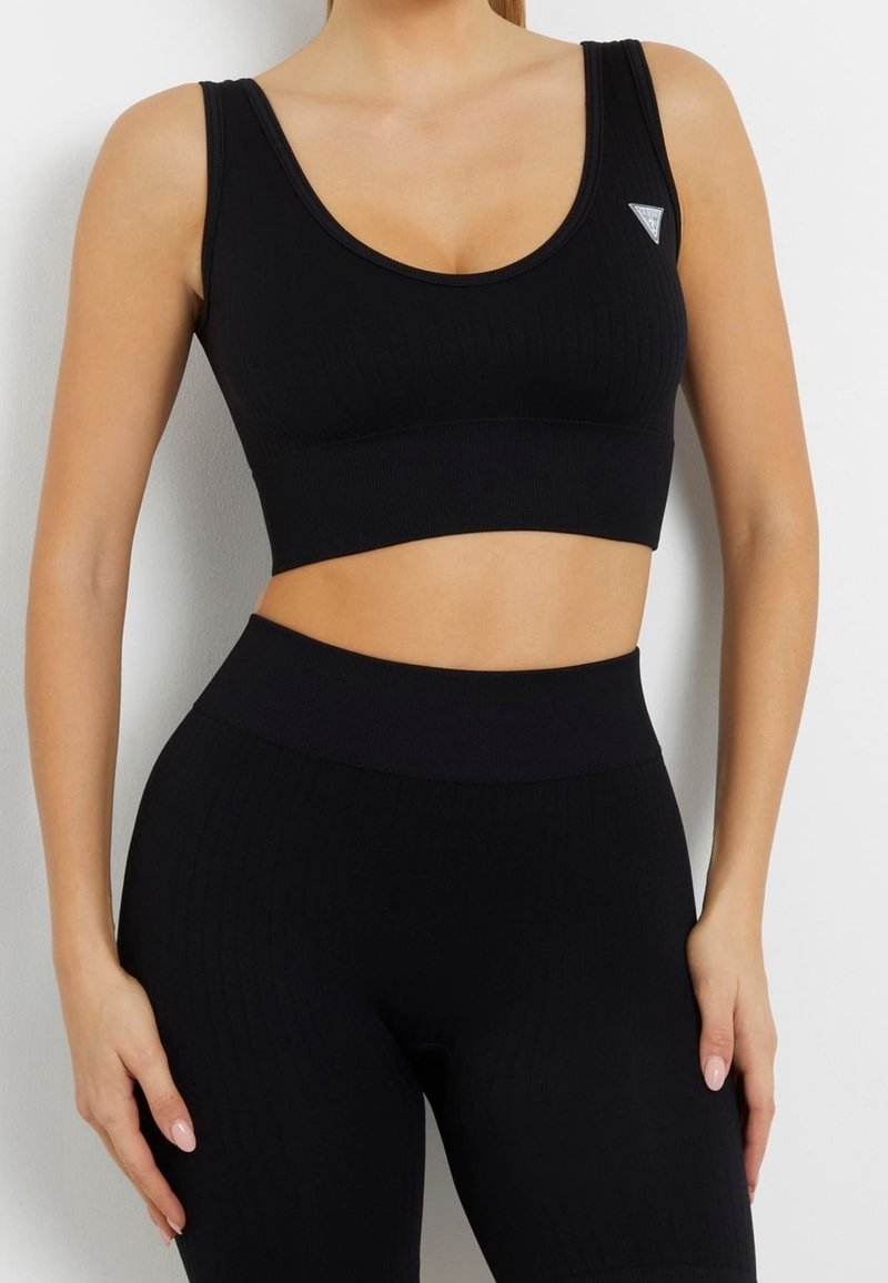 Žena v černé žebrované sportovní podprsence a odpovídajících kalhotách s vysokým pasem, stojící proti bílé zdi s rukama položenýma na stehnech.