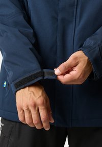 Navy jacket featuring a hook-and-loop cuff adjustment. Hands adjusting the cuff are visible. Material appears smooth and durable.