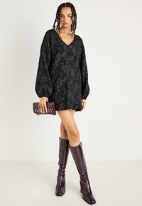 Melna ziedu kleita ar V veida kakla izgriezumu, pūkainām piedurknēm un brīvu piegriezumu. Apvienota ar augstiem, spīdīgiem, bordo zābakiem un atbilstošu mazu somiņu.