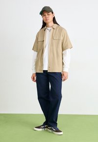 Chemise beige à manches courtes avec deux poches poitrine sur une chemise blanche à manches longues, assortie à un jean bleu foncé et des baskets multicolores.