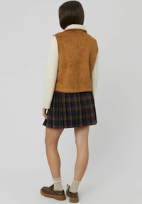 Gilet en suède synthétique marron avec un col en polaire beige, associé à une jupe à carreaux bleu marine avec des accents marron, complété par des chaussures et des chaussettes marron.