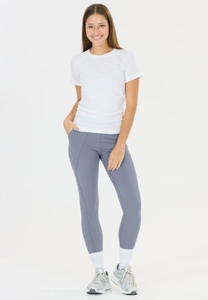 Lachende vrouw staand, gekleed in een wit shirt met korte mouwen, grijze leggings, witte sokken en grijze sneakers, met handen in de zakken, op een witte achtergrond.