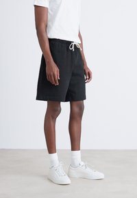 Black cotton shorts with an elastic waistband and drawstring, featuring side pockets and a relaxed fit, paired with white sneakers.