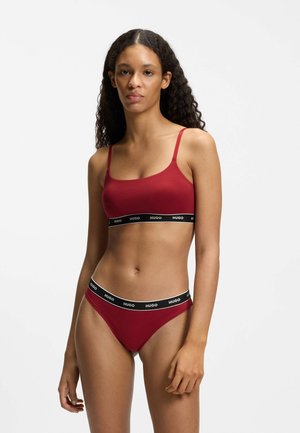 Frau mit langen, lockigen Haaren trägt ein rotes BH- und Slip-Set mit schwarzen elastischen Bändern, die mit "HUGO" gebrandmarkt sind, vor weißem Hintergrund.