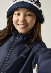 Niña sonriente con cabello largo y castaño lleva un sombrero tipo bucket azul marino con ribete blanco y una chaqueta con cremallera azul marino con el logo de Regatta en el pecho.