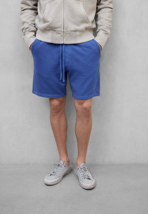 Person iført beige hettejakke med glidelås, blå shorts med snor, og grå joggesko, står mot en ensfarget grå vegg med hendene i lommene.