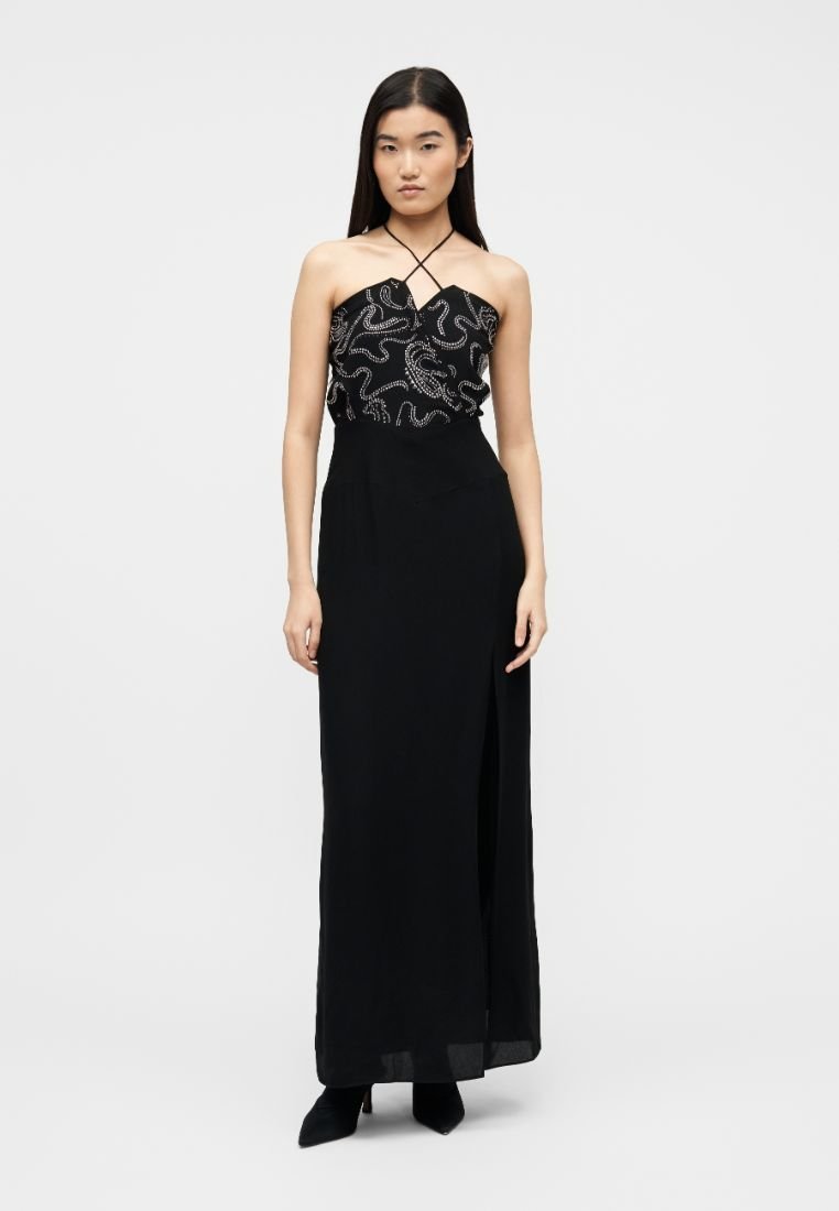Femme portant une longue robe noire bustier avec des motifs tourbillonnants blancs sur le corsage et une fente latérale, debout devant un fond uni.