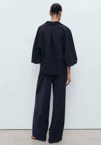 Černý oversize top s krátkými rukávy a záhybem na zadní straně, v kombinaci s volnými černými kalhotami. Hladká látka, minimalistický design.
