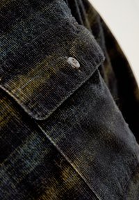 Primer plano de una chaqueta de pana negra, que muestra el acanalado vertical, un cierre de botones y sutiles destellos amarillos en la tela texturizada.