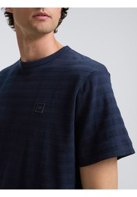 Camiseta texturizada azul marino con rayas ribeteadas horizontales y un pequeño parche de logotipo negro en el lado izquierdo del pecho.