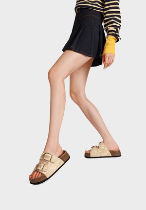 Persona che indossa una gonna plissettata nera, una maglietta a righe a maniche lunghe e sandali beige con due fibbie che cammina su uno sfondo semplice.