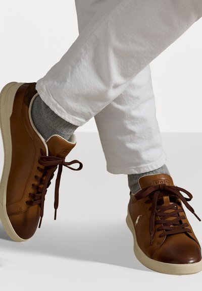 Sneaker in pelle marrone con dettagli in marrone scuro, stringhe piatte e suole beige, indossati con calzini grigi e pantaloni bianchi.