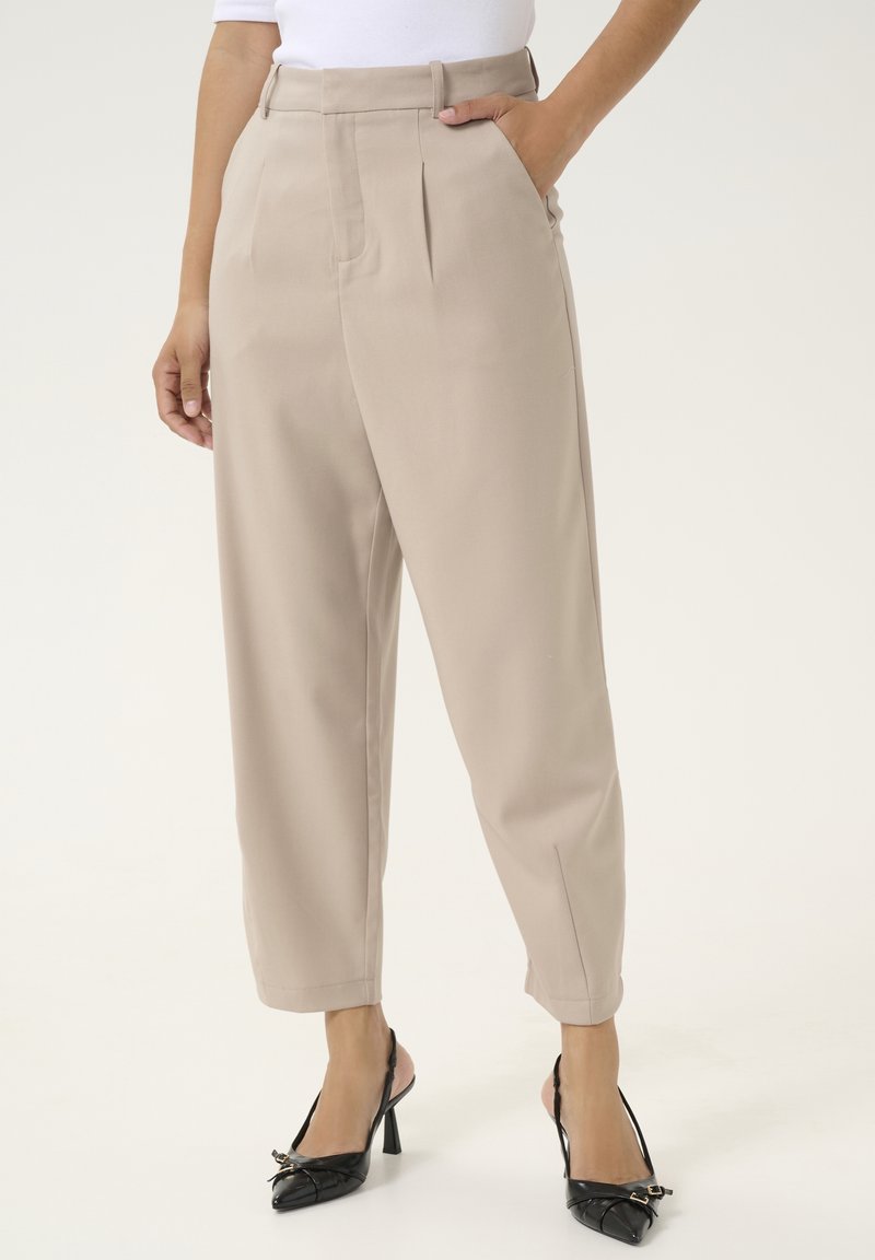 Pantalon beige taille haute fuselé avec plis, porté avec des talons slingback noirs à bout pointu et un haut blanc, main dans la poche.