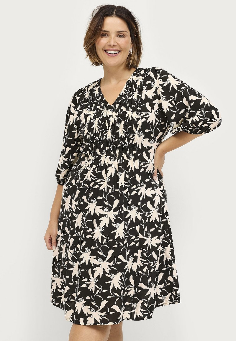 Schwarzes kurzärmliges Kleid mit einem floralen Muster in Creme, mit einem gerüschten Oberteil und V-Ausschnitt, das oberhalb des Knies endet.