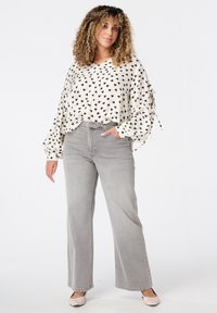 Femme aux cheveux bouclés portant une blouse blanche avec des cœurs noirs, un jean large taille haute gris et des chaussures beiges, se tenant avec une main dans la poche.