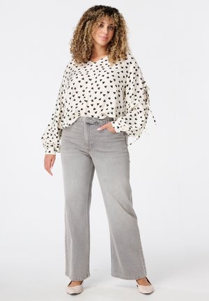 Femme aux cheveux bouclés portant une blouse blanche avec des cœurs noirs, un jean large taille haute gris et des chaussures beiges, se tenant avec une main dans la poche.