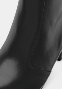 Detailný záber lesklých čiernych kožených členkových topánok s viditeľným šitím a blokovým podpätkom na bielom pozadí.